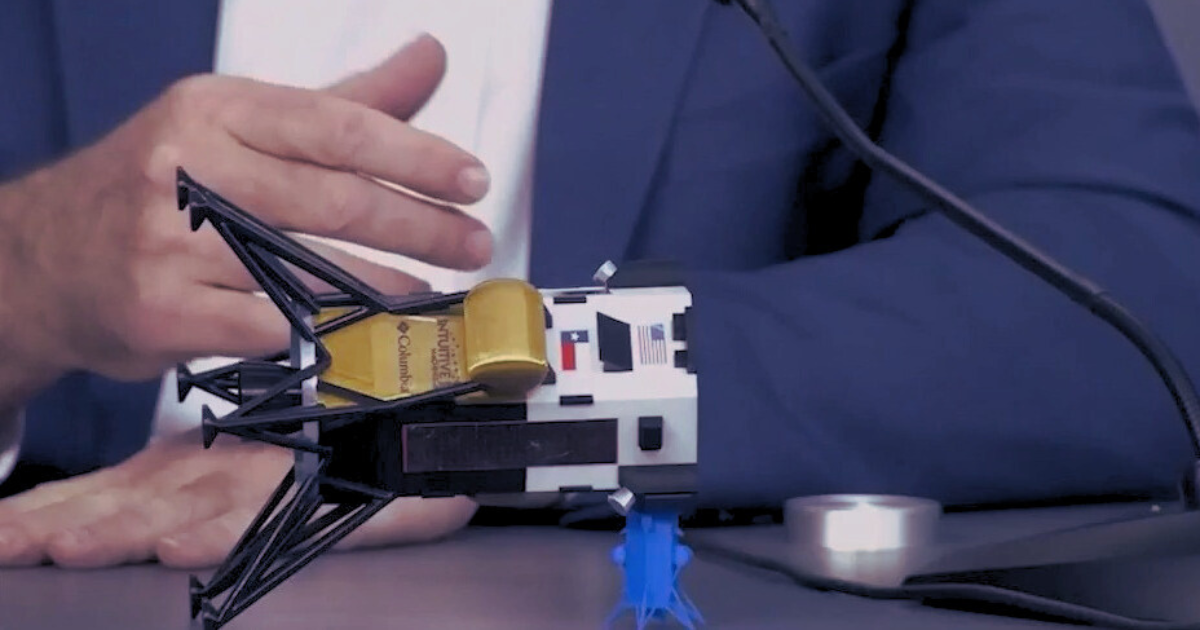 A close-up of a model Odysseus Moon lander on a desk, with Intuitive Machines CEO Stephen Altemus' hand and torso behind it.