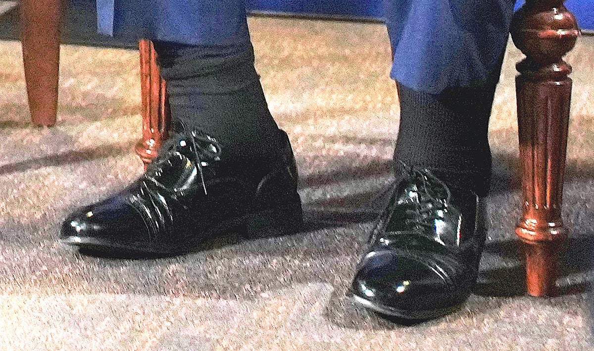 A zoomed-in, color-corrected photo of president Donald Trump's ankles, which appear swollen. He wears navy blue pants, black socks, and shiny black Oxford shoes with his feet on a grey carpet. 