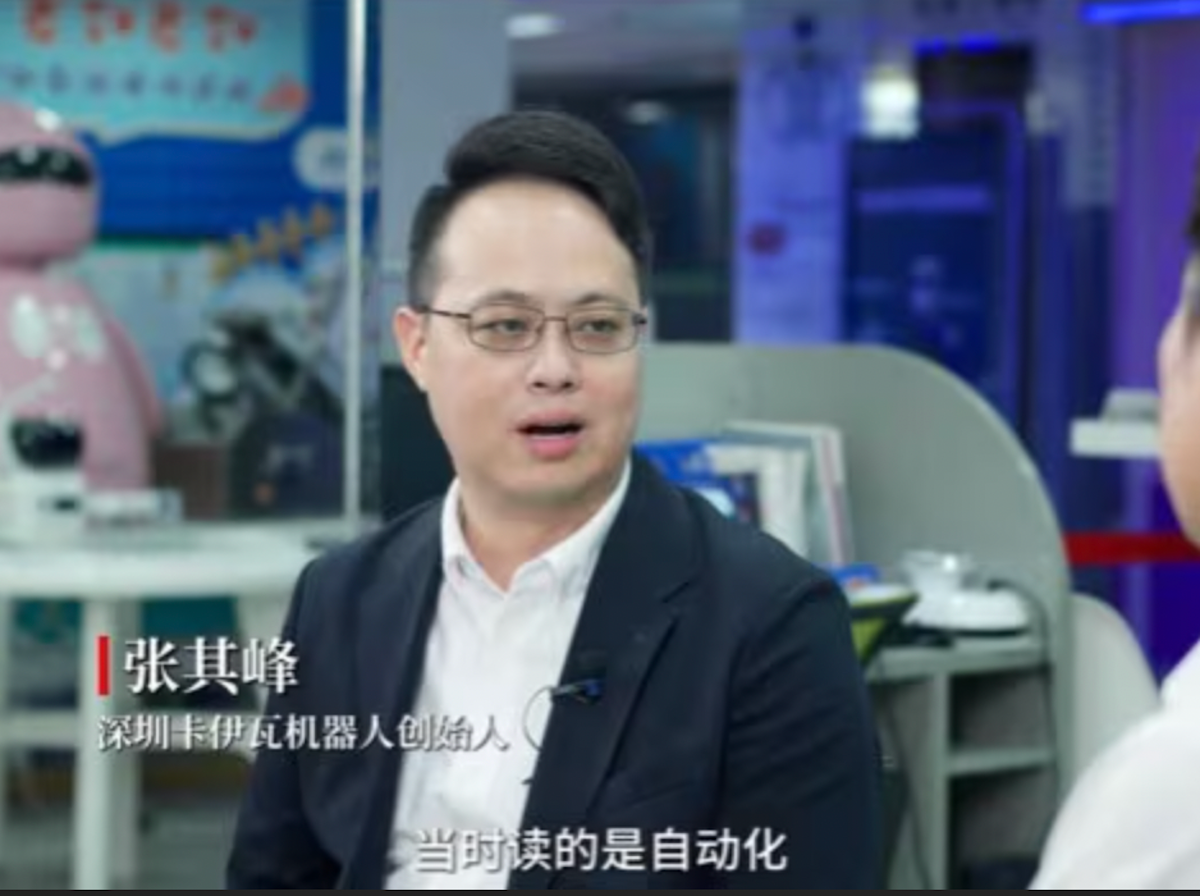 An Asian man with round, wire-rimmed glasses looks at someone off camera in what appears to be a newsroom, which is bathed in blue and chrome. There is white text over the top of the image, written in Mandarin.